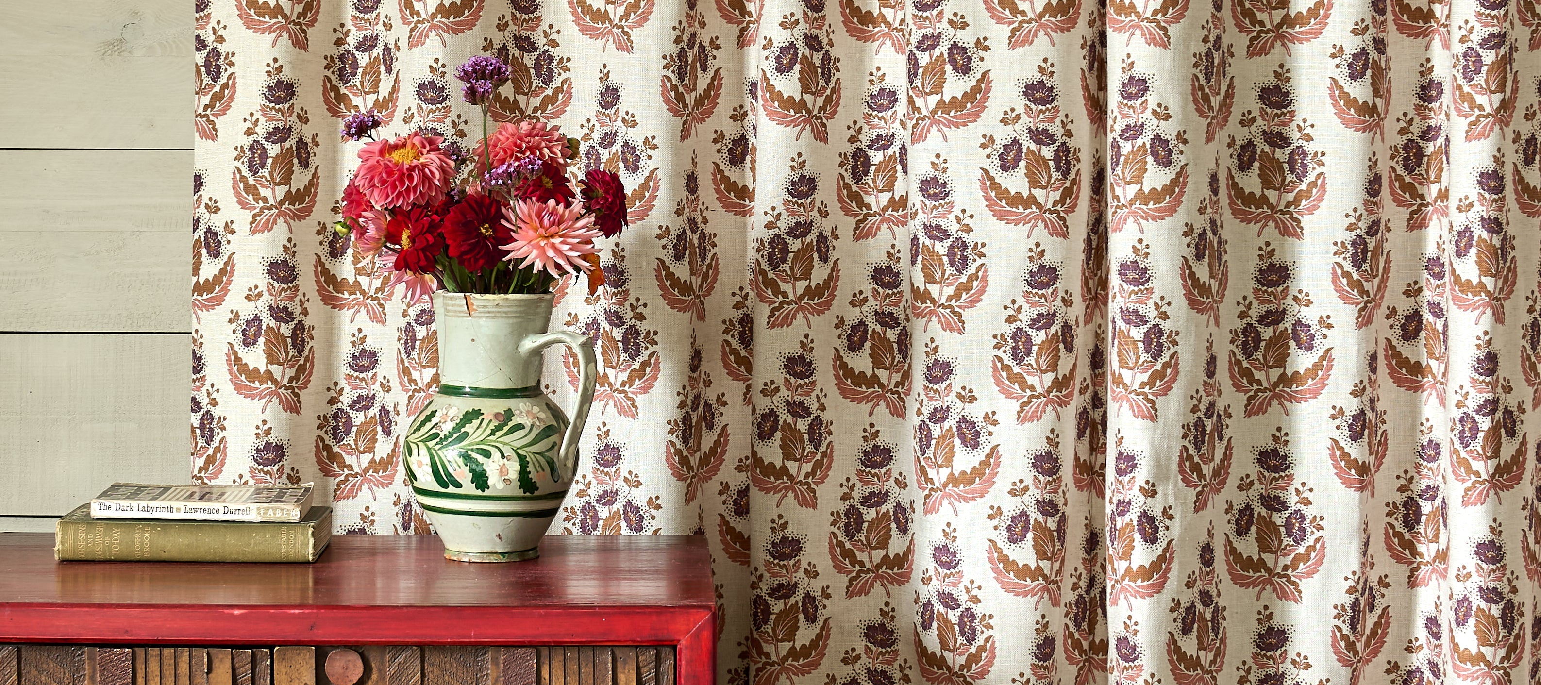 A table with fabric swatches sits in front of a rack of Rtolls of printed fabric, in the background we see two women screen printing at table. 