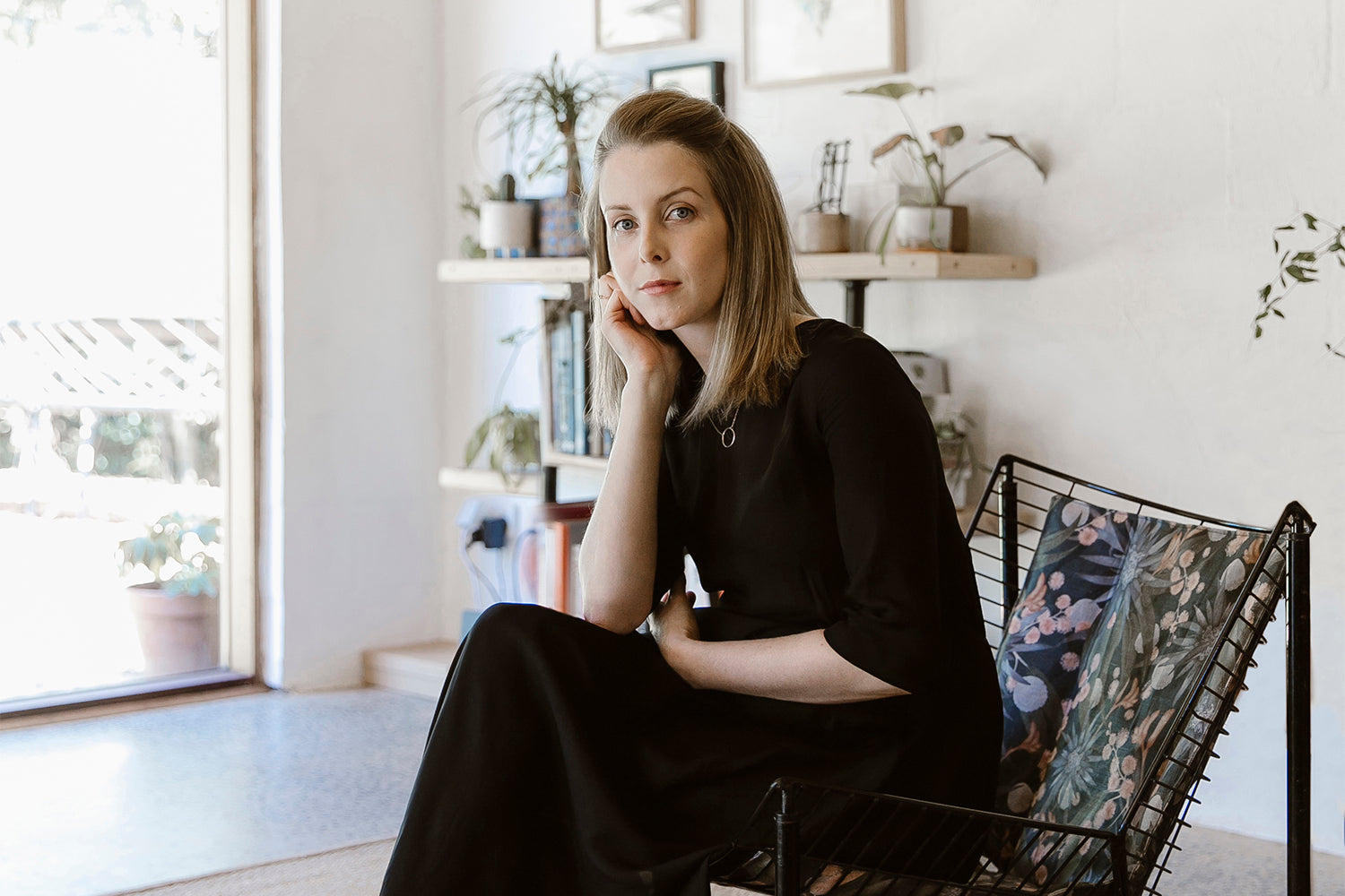 A blonde woman wearing a long black dress seated in a low-slung black armchair, looking at the camera and resting her head on her hand.