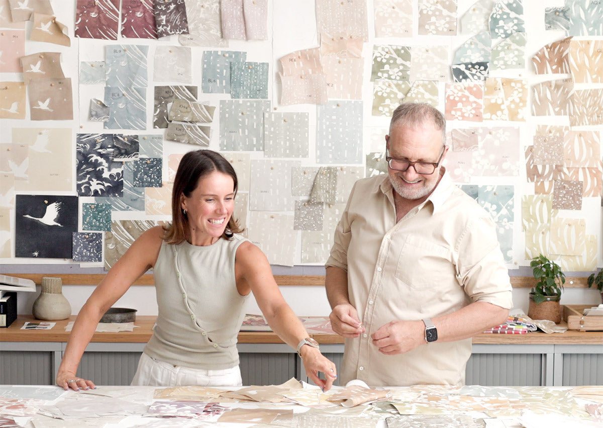 Black and white three-quarter shot of a smiling man with glasses, standing with arms crossed in front of wallpaper in a bird print.