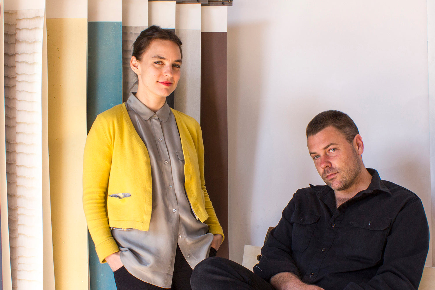 A seated man dressed in all black and a standing woman next to him, both in front of a rack of large hanging wallpaper swatches.