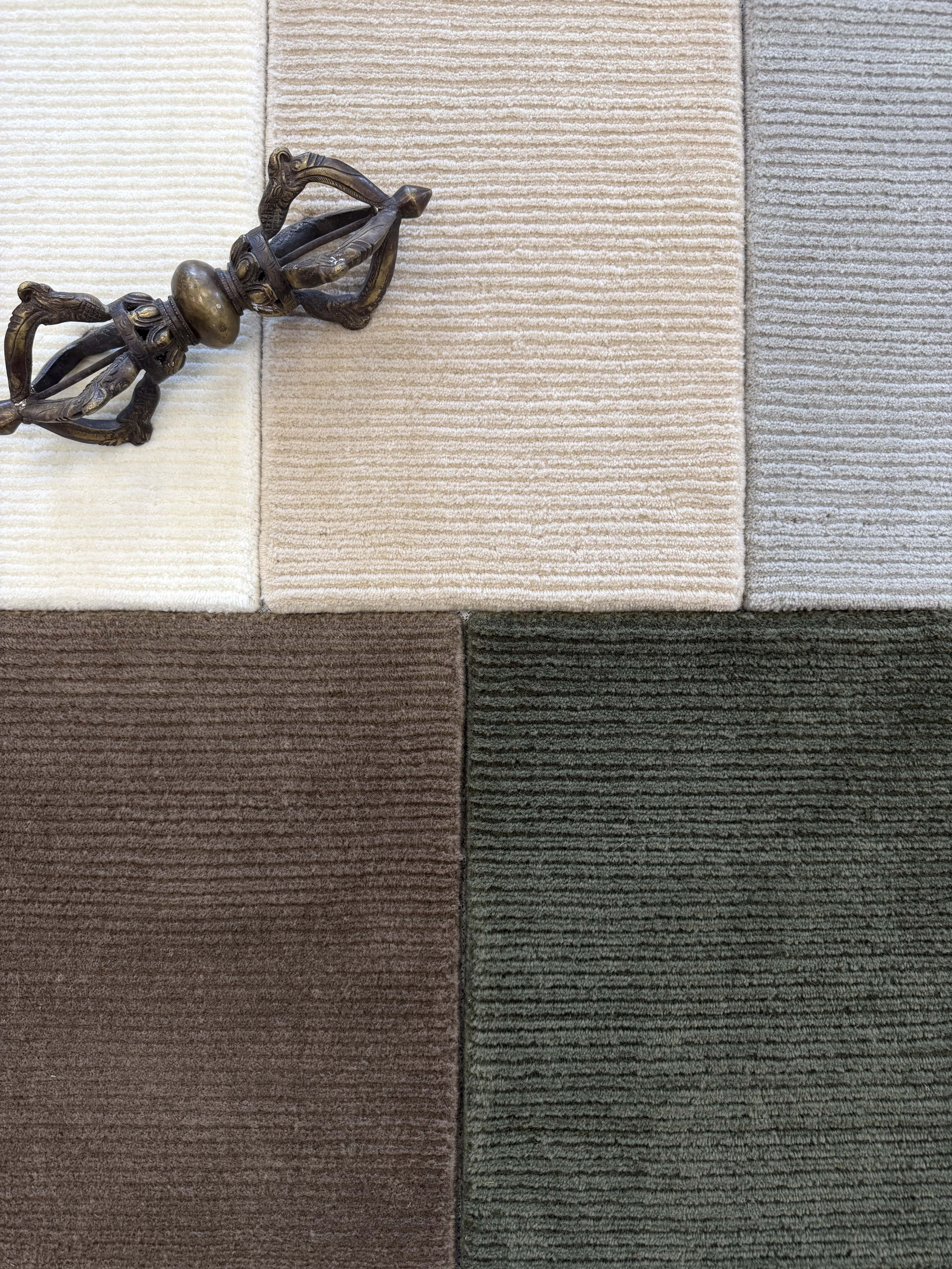 Close up of broadloom carpet samples with solid cut pile ribbed stripe in white, beige, grey brown and green with an ornamental brass sculpture. 