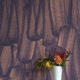 A vase of flowers stands in front of a wall papered in an abstract paint print in dark grey on a purple field.