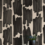 A vase of flowers stands in front of a wall papered in an abstract paint swatch print in black on a cream field.