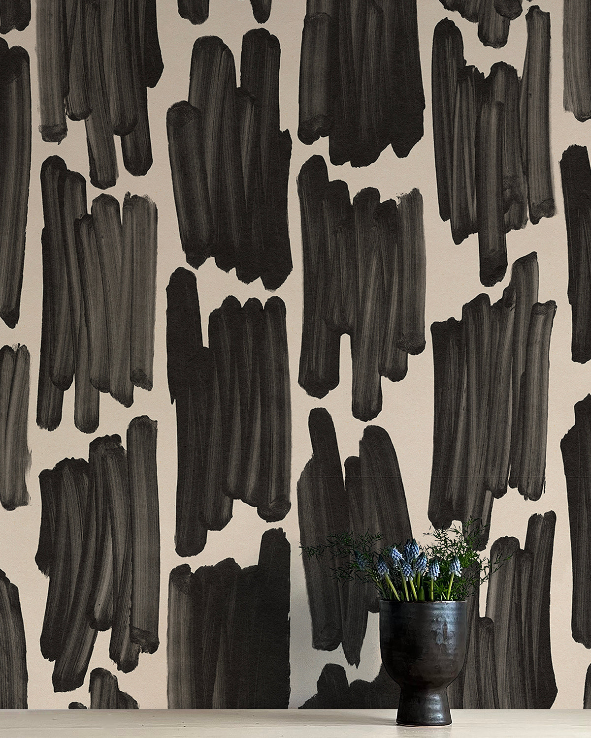 A vase of flowers stands in front of a wall papered in an abstract paint swatch print in black on a cream field.