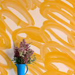 A vase of flowers stands in front of a wall covered in an abstract paintstroke print in orange on a cream field.