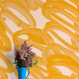 A vase of flowers stands in front of a wall covered in an abstract paintstroke print in orange on a cream field.