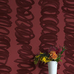 A vase of flowers stands in front of a wall covered in an abstract scribble print in black on a red field.