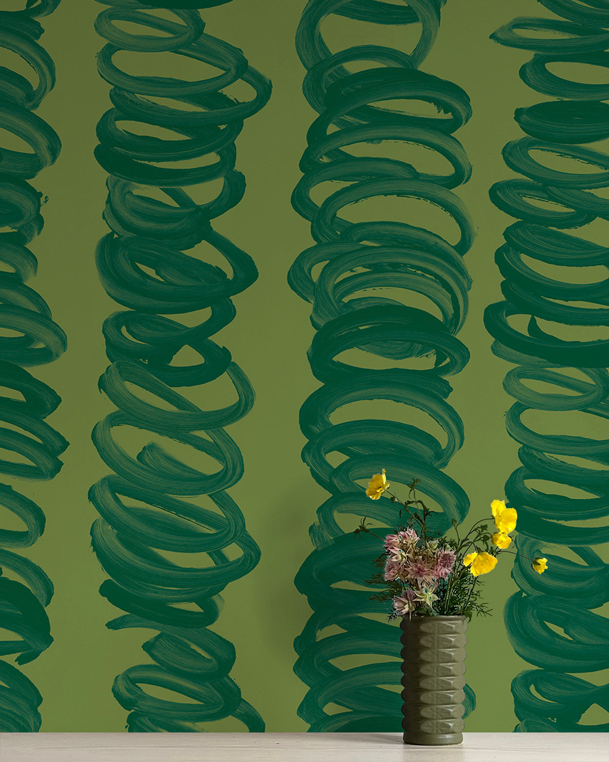 A vase of flowers stands in front of a wall covered in an abstract scribble print in green on an olive field.