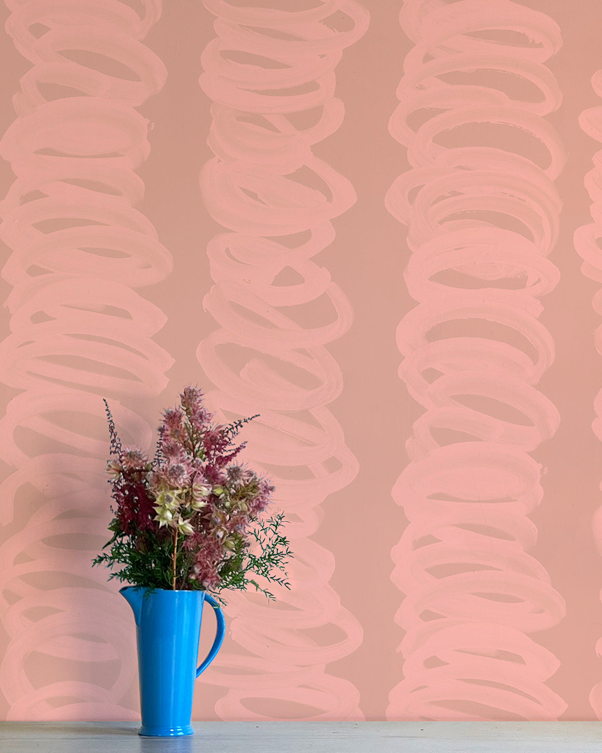 A vase of flowers stands in front of a wall covered in an abstract scribble print in light pink on a tan field.