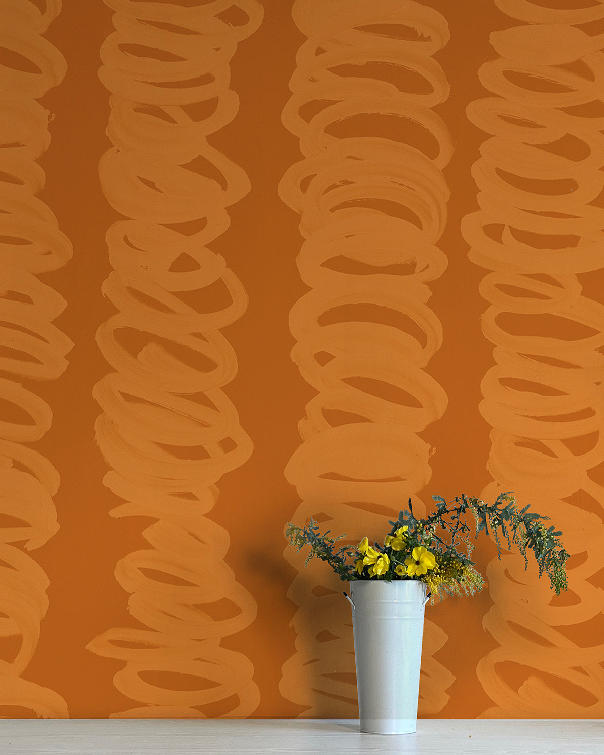 A vase of flowers stands in front of a wall covered in an abstract scribble print in orange on a rust field.