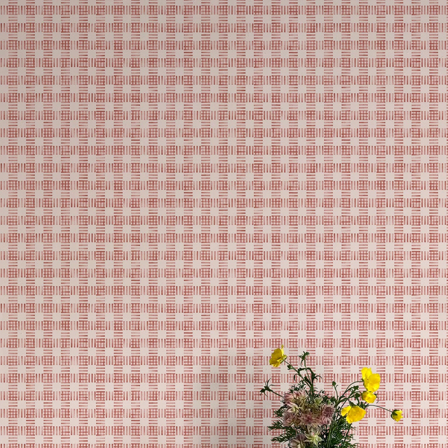 A vase of flowers stands in front of a wall papered in a dense plaid pattern in shades of cream and red.