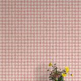 A vase of flowers stands in front of a wall papered in a dense plaid pattern in shades of cream and red.