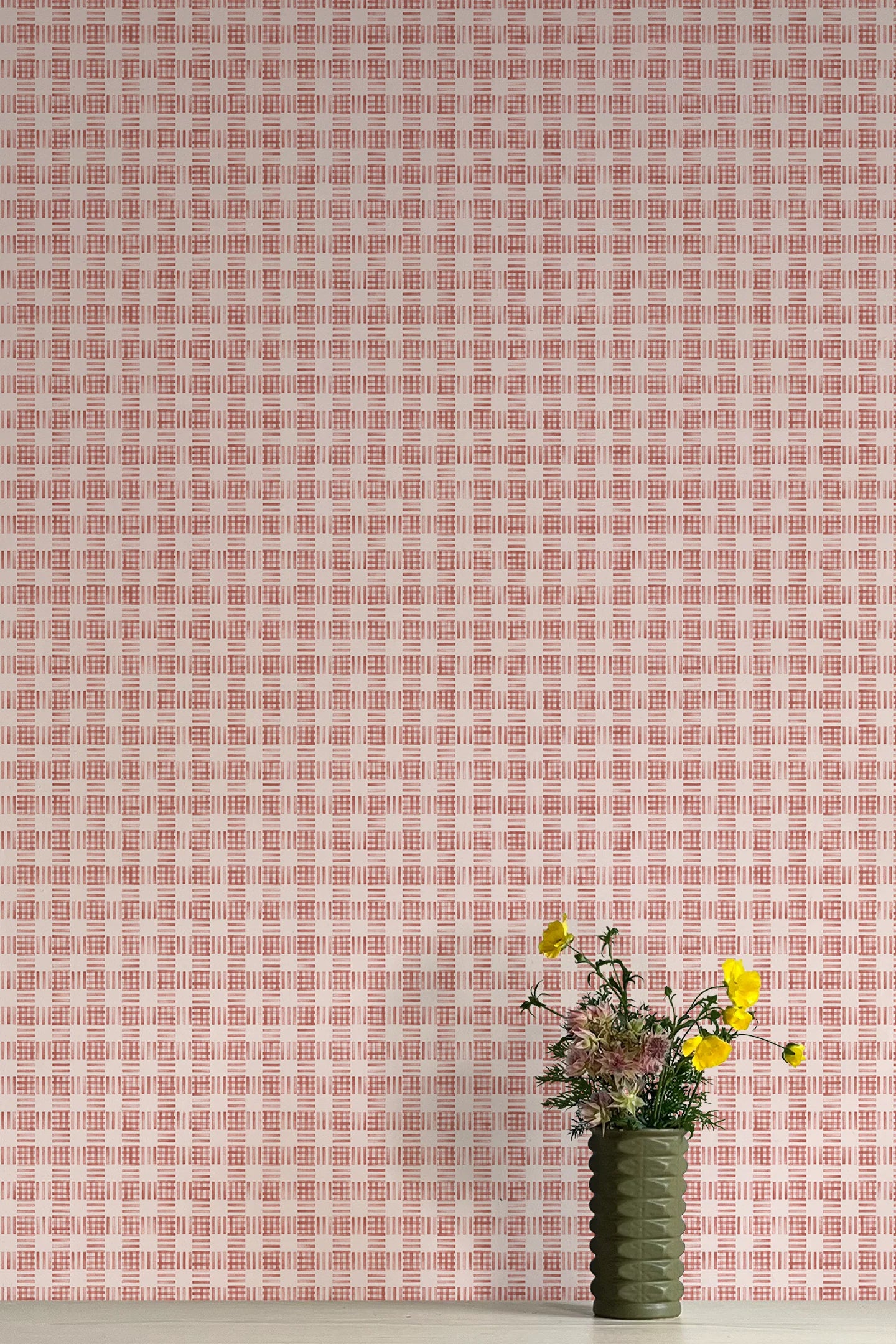 A vase of flowers stands in front of a wall papered in a dense plaid pattern in shades of cream and red.