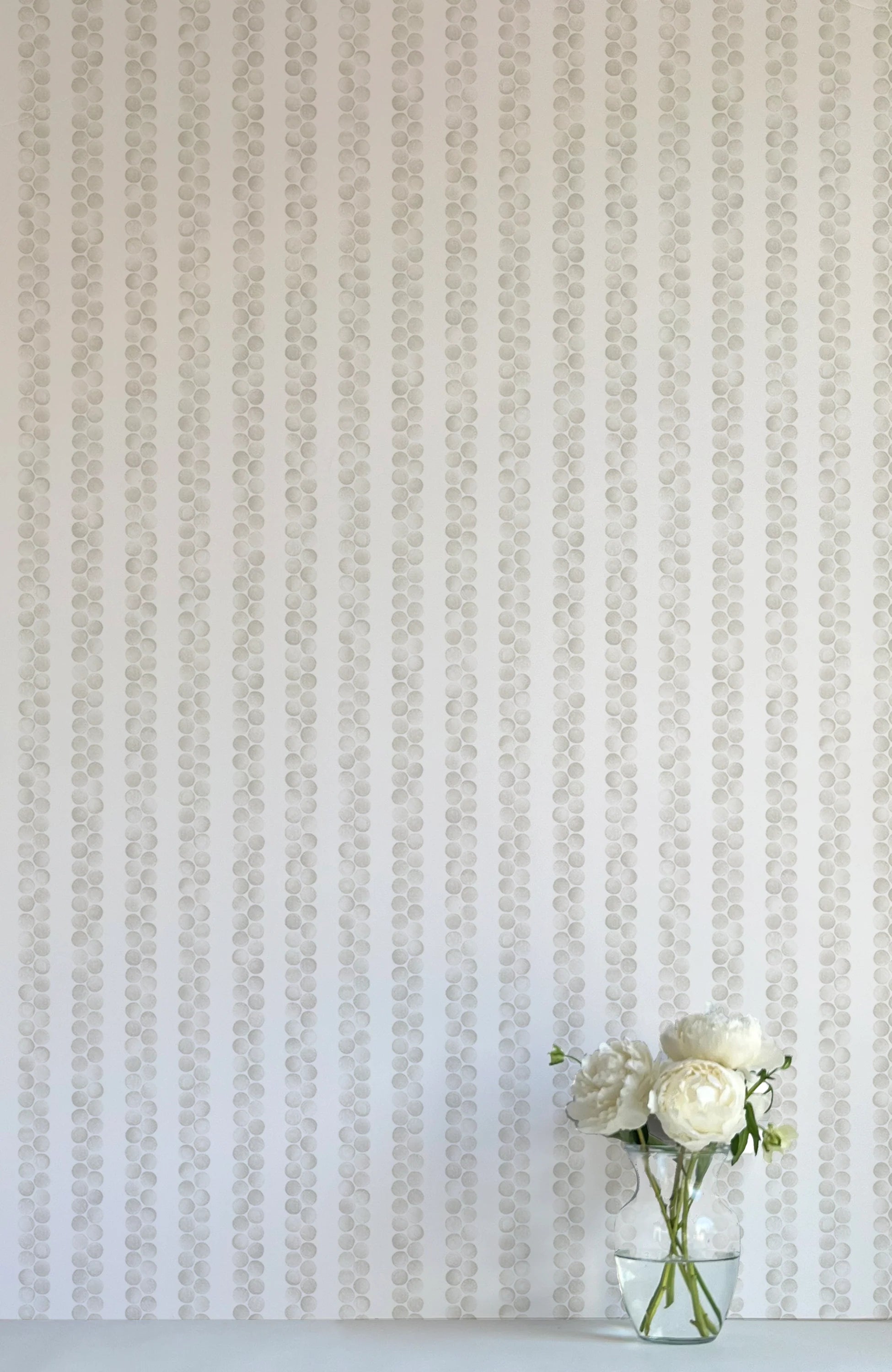 A vase of flowers stands in front of a wall covered in a repeating striped circular print in gray on a light gray field.