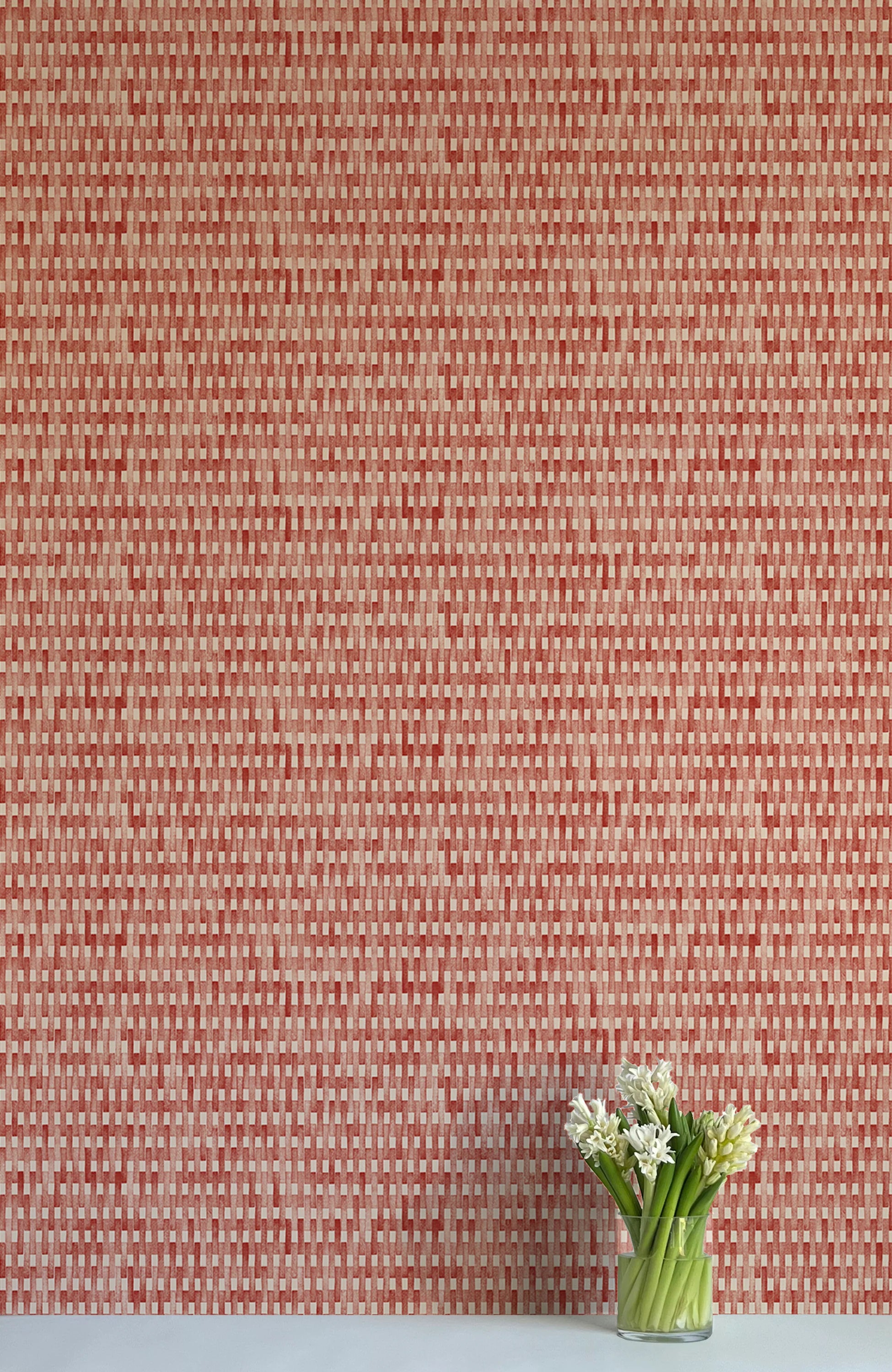 A vase of flowers stands in front of a wall covered in a textural grid print in shades of red and cream.