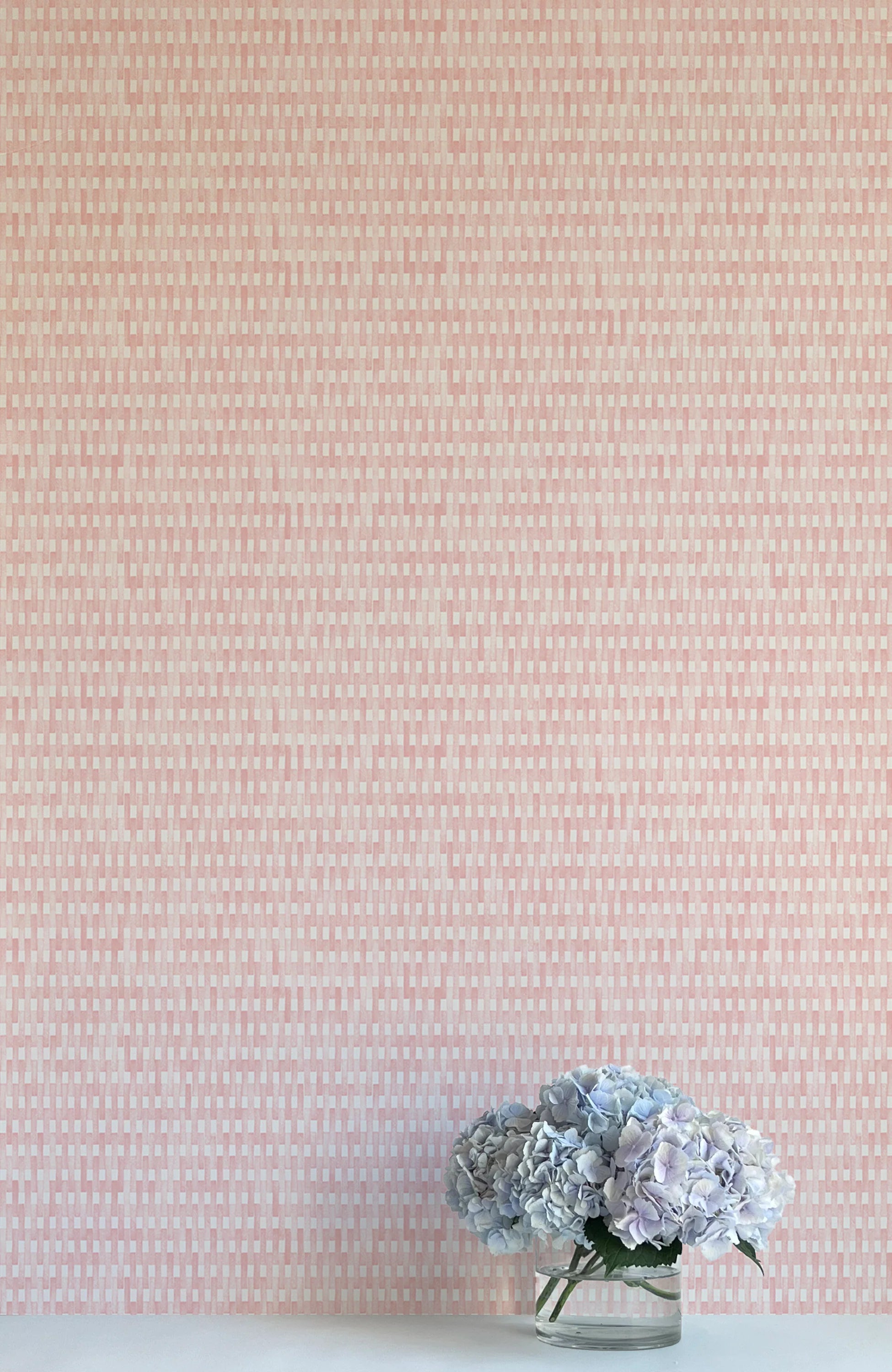 A vase of flowers stands in front of a wall covered in a textural grid print in shades of pink and cream.
