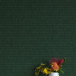 A vase of flowers stands in front of a wall covered in a textural grid print in shades of dark green.