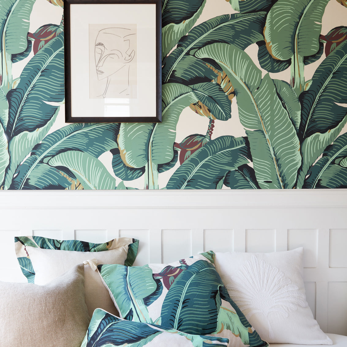 A styled living room scene featuring a white ottoman  with tropical banana leaf patterned pillows and matching wall wallpaper. A framed minimalist line art print hangs on the wall.