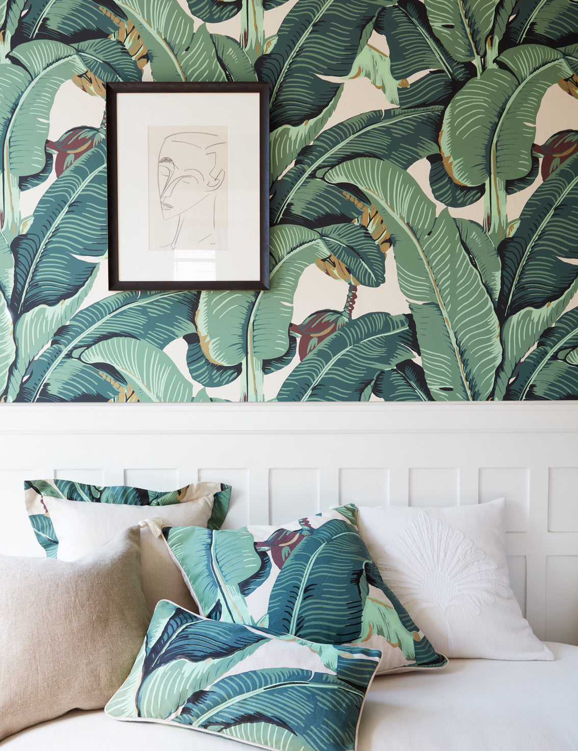 A styled living room scene featuring a white ottoman  with tropical banana leaf patterned pillows and matching wall wallpaper. A framed minimalist line art print hangs on the wall.