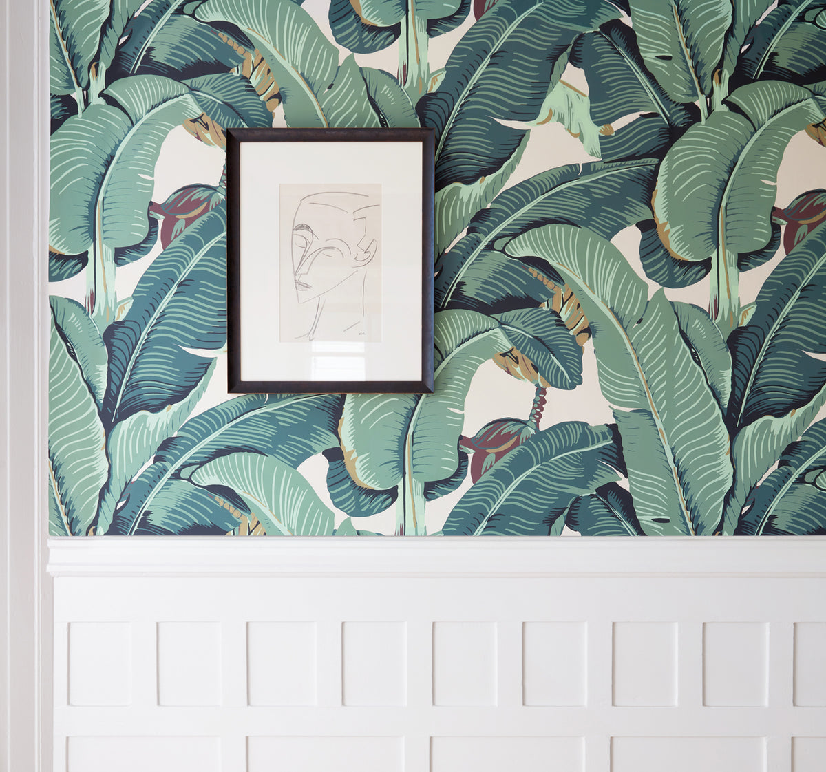 Tropical banana leaf wallpaper in shades of green and teal on a cream background, styled on a wall above a white wainscoted panel with a small framed abstract print.