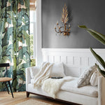 A styled living room corner featuring a white sofa with cream and black patterned pillows, a white throw blanket, a gold wall sconce, a dark gray wall with white wainscoting, and tropical leaf patterned curtains.