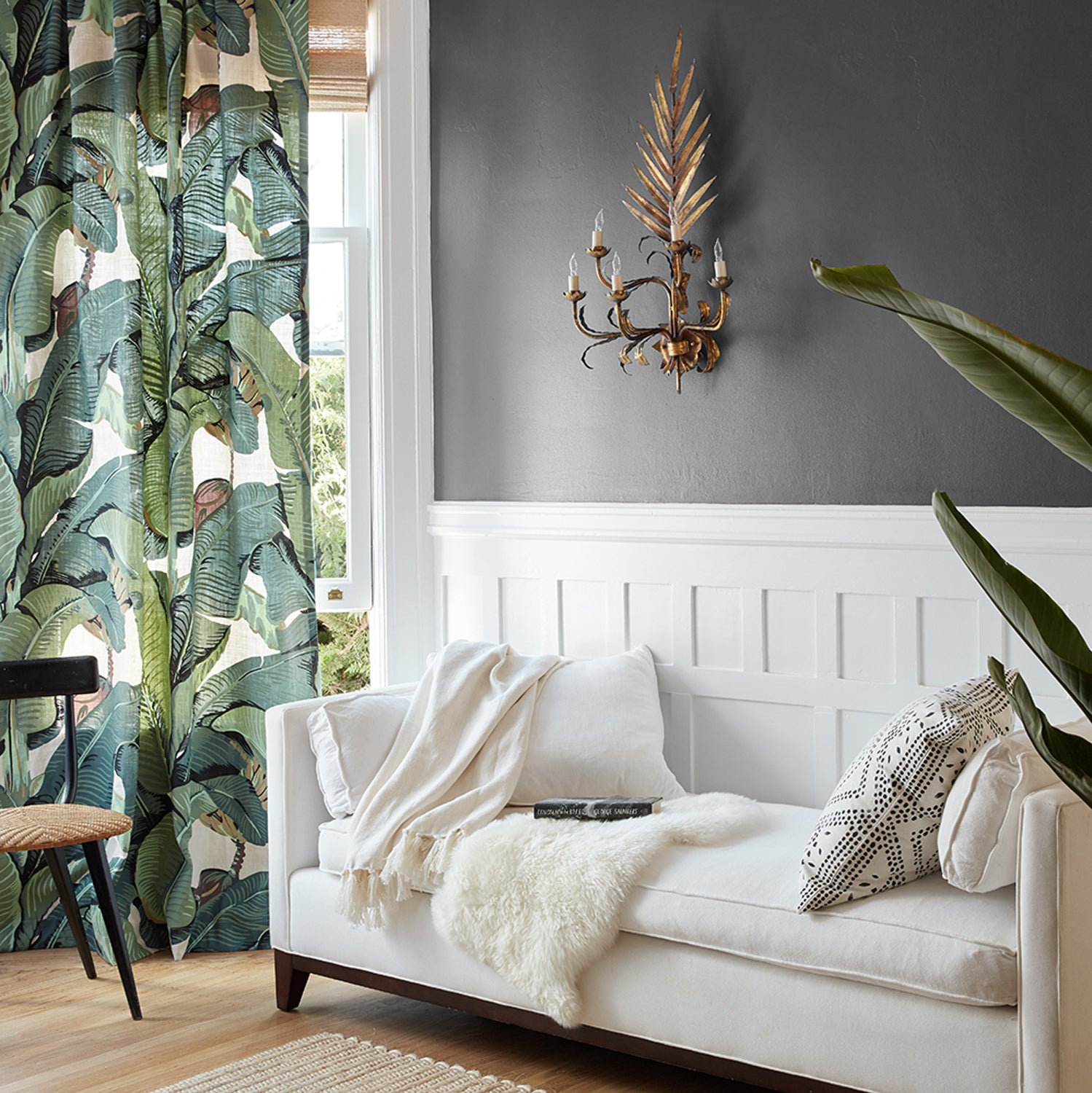 A styled living room corner featuring a white sofa with cream and black patterned pillows, a white throw blanket, a gold wall sconce, a dark gray wall with white wainscoting, and tropical leaf patterned curtains.