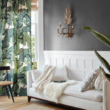 A styled living room corner featuring a white sofa with cream and black patterned pillows, a white throw blanket, a gold wall sconce, a dark gray wall with white wainscoting, and tropical leaf patterned curtains.