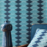 A close-up of a dark navy wallpaper with a repeating light blue geometric diamond pattern, styled in a room setting with a matching blue chair, a blue ceramic vase, and a patterned pillow.