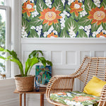 A styled interior corner featuring a bold tropical floral wallpaper with orange, white, and green leaves, a wicker armchair with a matching cushion, a wooden side table with a potted plant, and white wainscoting.
