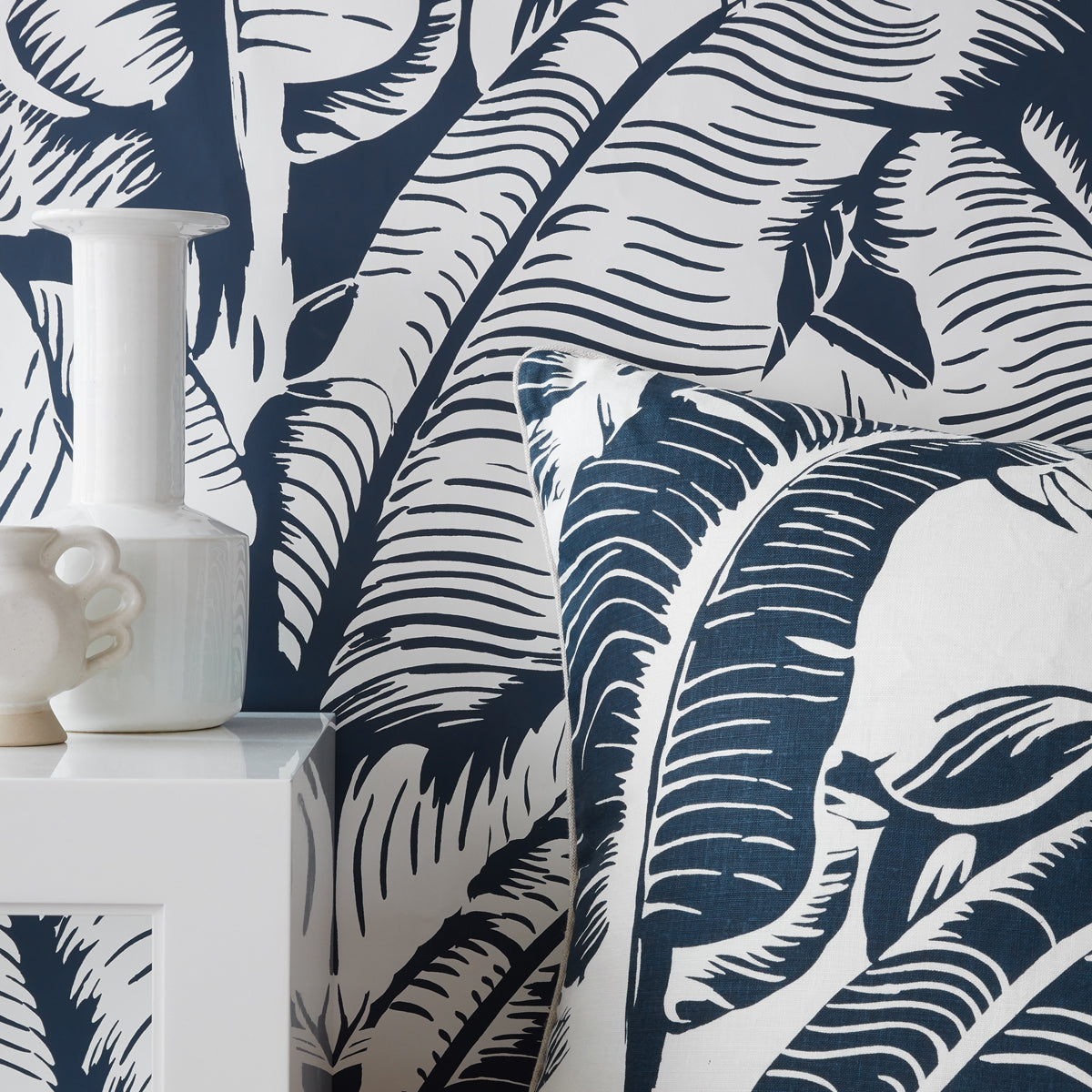 A close-up of a tropical leaf patterned wallpaper and matching throw pillow in navy blue and white, styled with a white ceramic vase on a white side table.