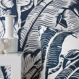 A close-up of a tropical leaf patterned wallpaper and matching throw pillow in navy blue and white, styled with a white ceramic vase on a white side table.