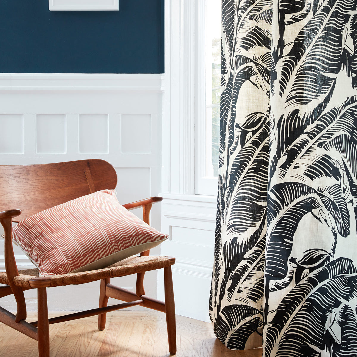 A close-up of a curtain panel featuring a bold black and cream tropical leaf pattern, hanging next to a window in a styled room with a wooden chair and a pink cushion.