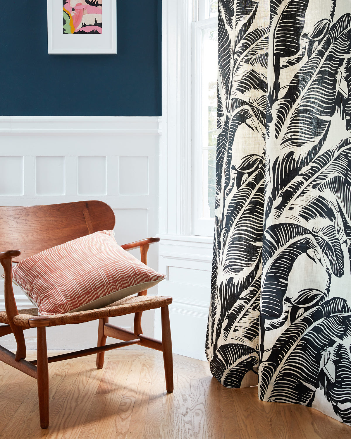 A close-up of a curtain panel featuring a bold black and cream tropical leaf pattern, hanging next to a window in a styled room with a wooden chair and a pink cushion.