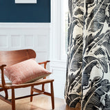 A close-up of a curtain panel featuring a bold black and cream tropical leaf pattern, hanging next to a window in a styled room with a wooden chair and a pink cushion.