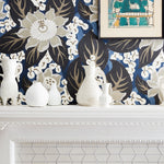 A styled interior scene showing a white fireplace mantel decorated with white ceramic vases, set against a bold floral wallpaper in navy, cream, and taupe. A framed art print hangs on the wall above the mantel.