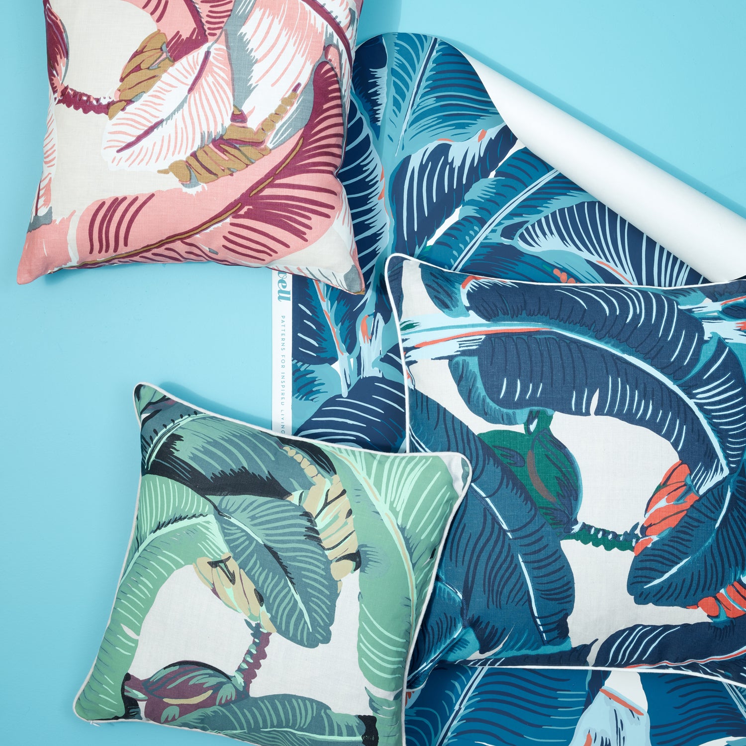 A styled flat-lay of tropical leaf patterned home textiles on a bright blue background, including two decorative throw pillows and a matching rolled fabric panel.