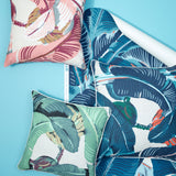 A styled flat-lay of tropical leaf patterned home textiles on a bright blue background, including two decorative throw pillows and a matching rolled fabric panel.
