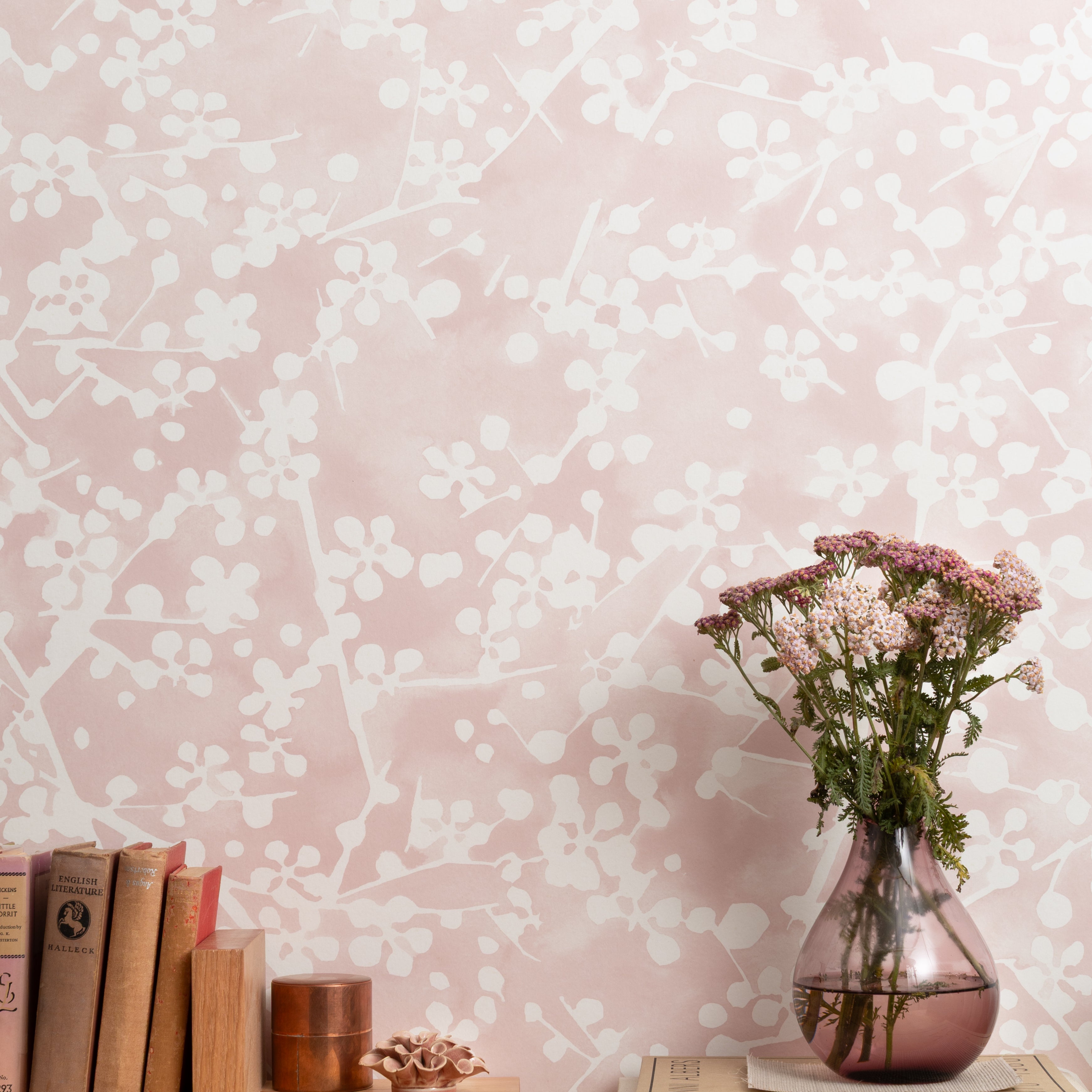 Mantle vignette with vase and flowers and antique books in front of a wallpapered in painterly branch print in white on a pale pink field.