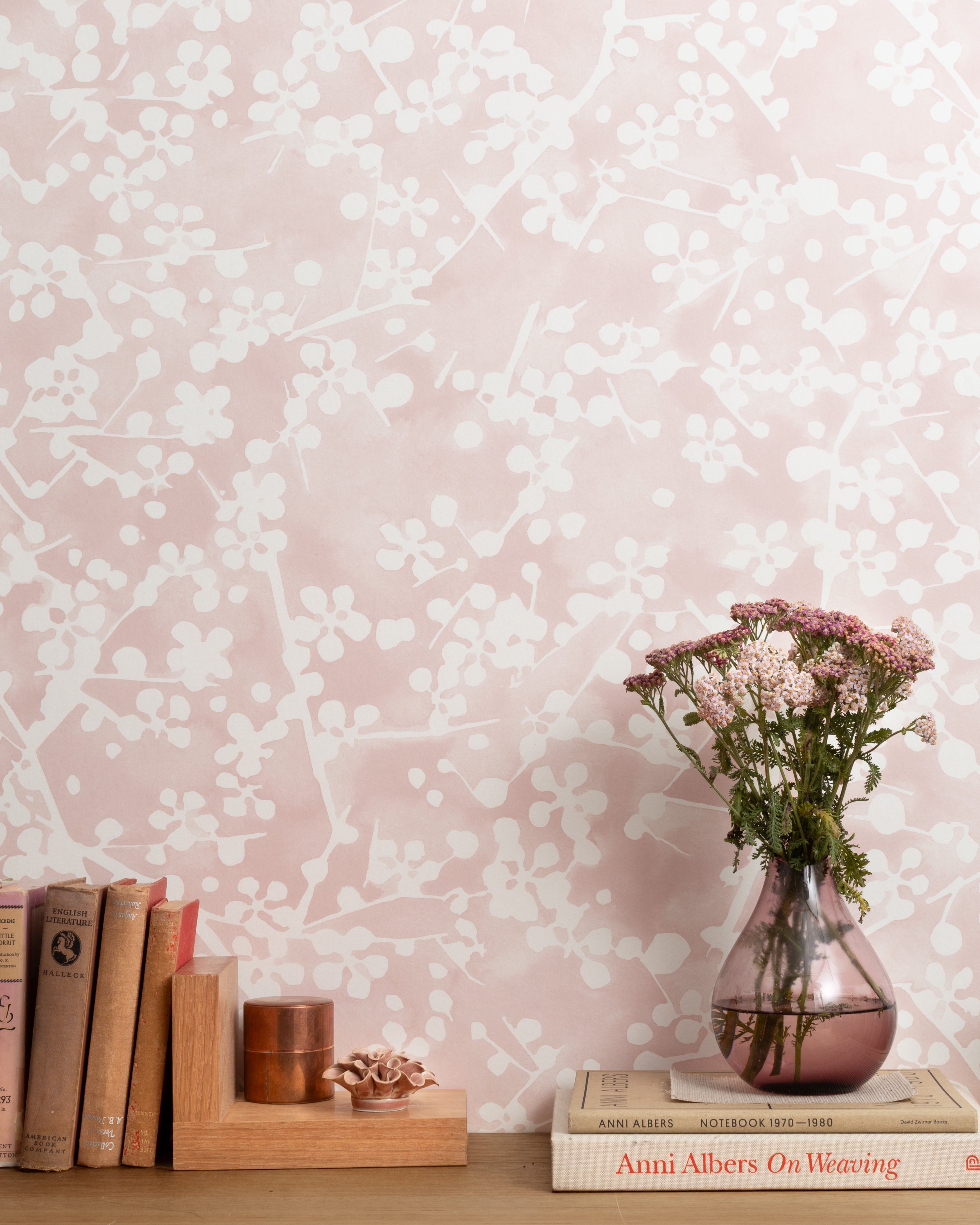 Mantle vignette with vase and flowers and antique books in front of a wallpapered in painterly branch print in white on a pale pink field.