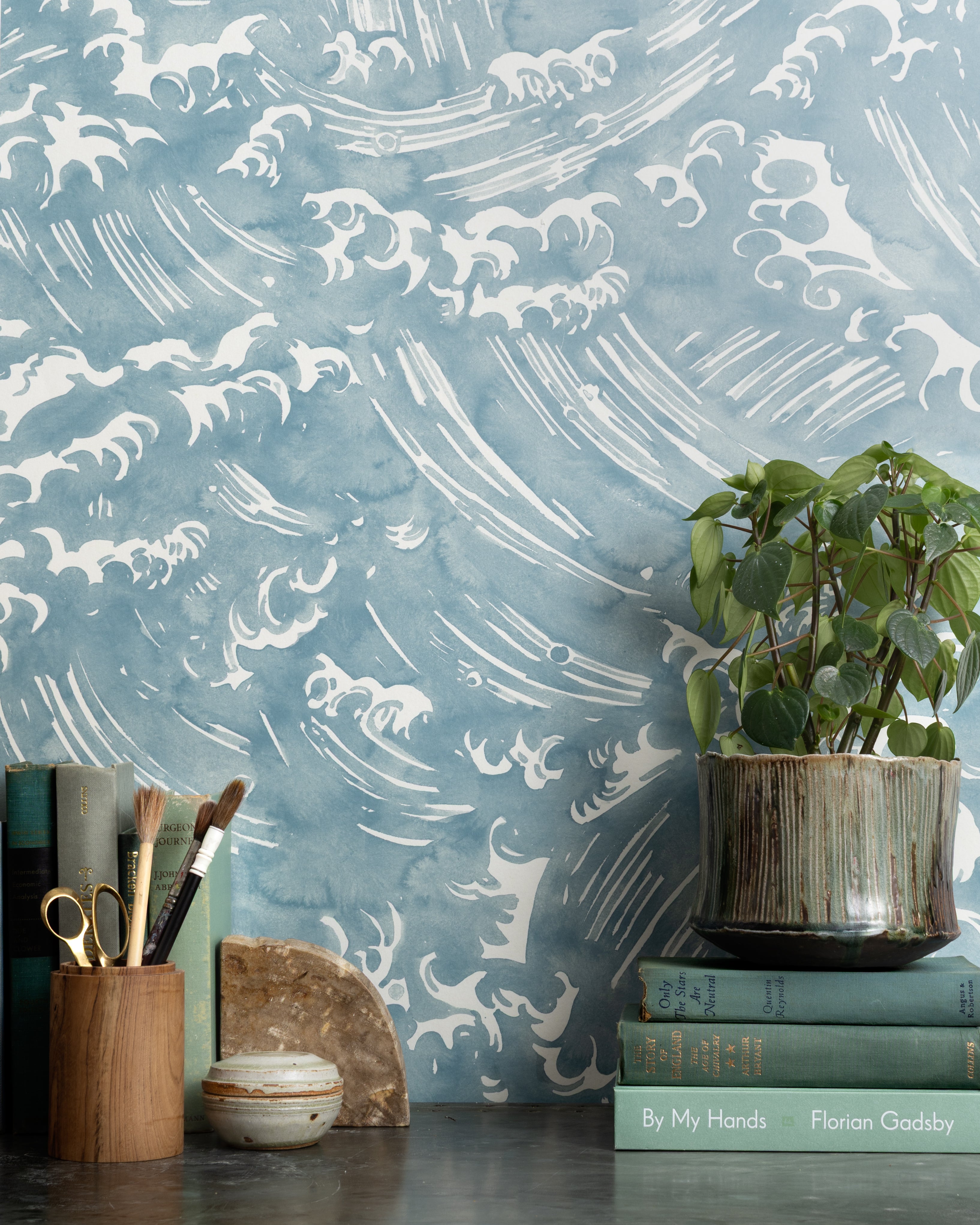 Mantle vignette with antique books, potted plant and pencil jar against wallpaper  in a playful wave print in white on a chambray blue field.