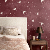 Bedroom scene with layered printed bed clothes in shades of taupe and pink against a wallpaper with stars and flying geese in white on a crimson watercolor field.