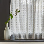 Detail of a window sill with a vase with a branch against a curtain in sheer fabric in a check pattern in ivory.