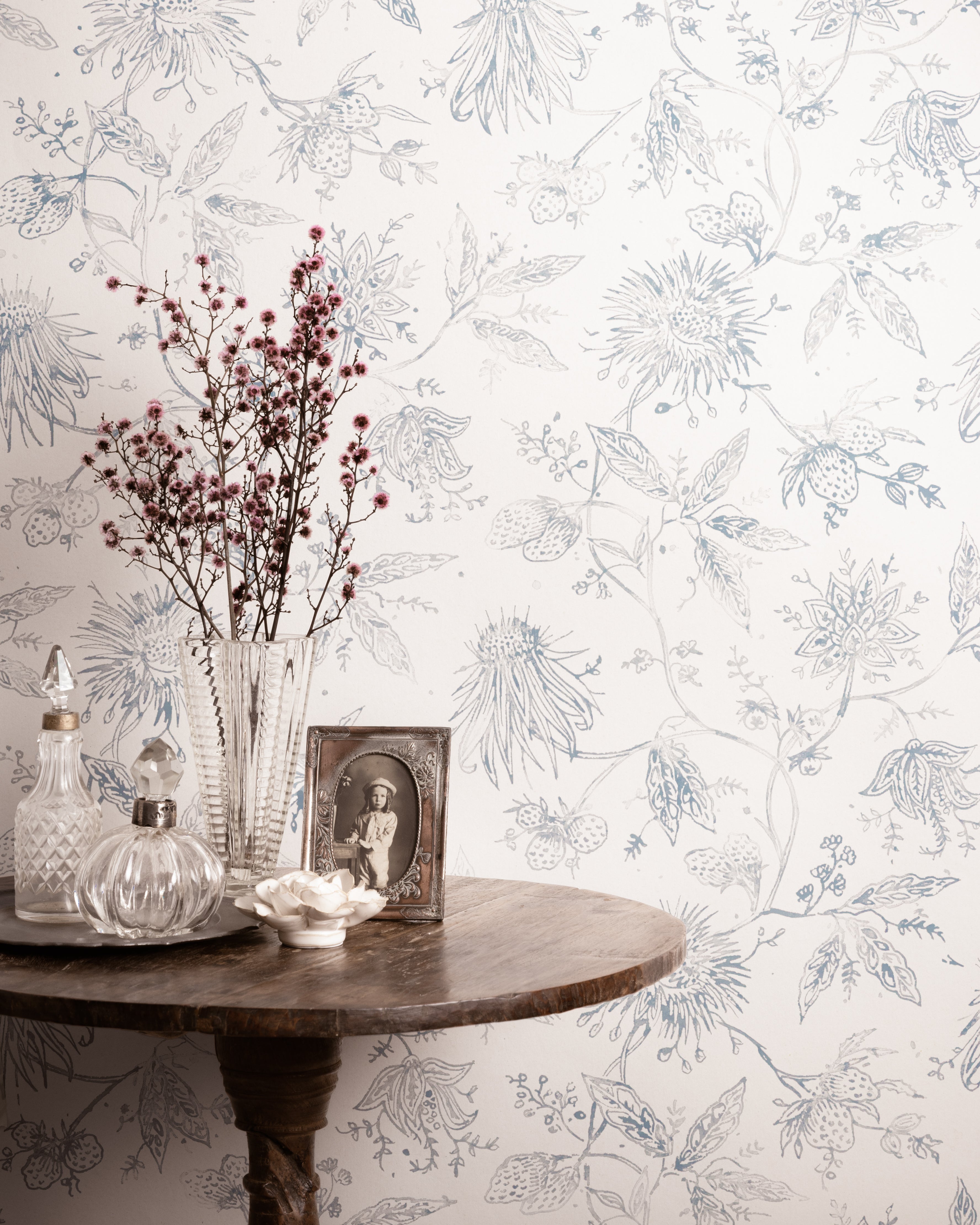 Decorative table with a vase, bottles, and photo frame against a floral wallpapered wall.