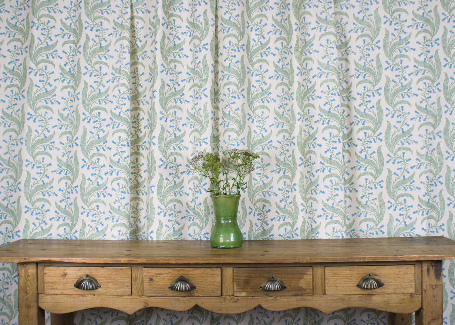 Wooden console table with vase and flowers against curtain in a scrolling floral pattern in blue and green on a cream field. 