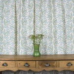 Wooden console table with vase and flowers against curtain in a scrolling floral pattern in blue and green on a cream field. 