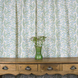 Wooden console table with vase and flowers against curtain in a scrolling floral pattern in blue and green on a cream field. 