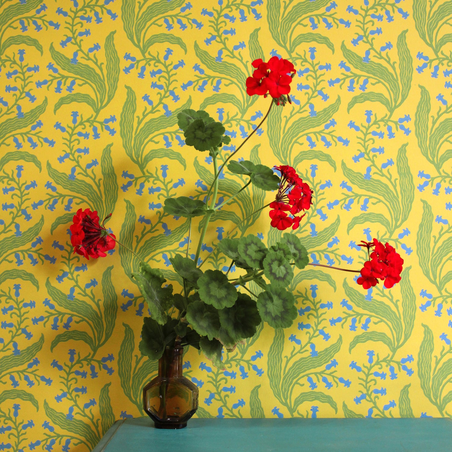 Teal table with vase and flowers against wallpaper in a scrolling floral pattern in blue and green on a yellow field. 