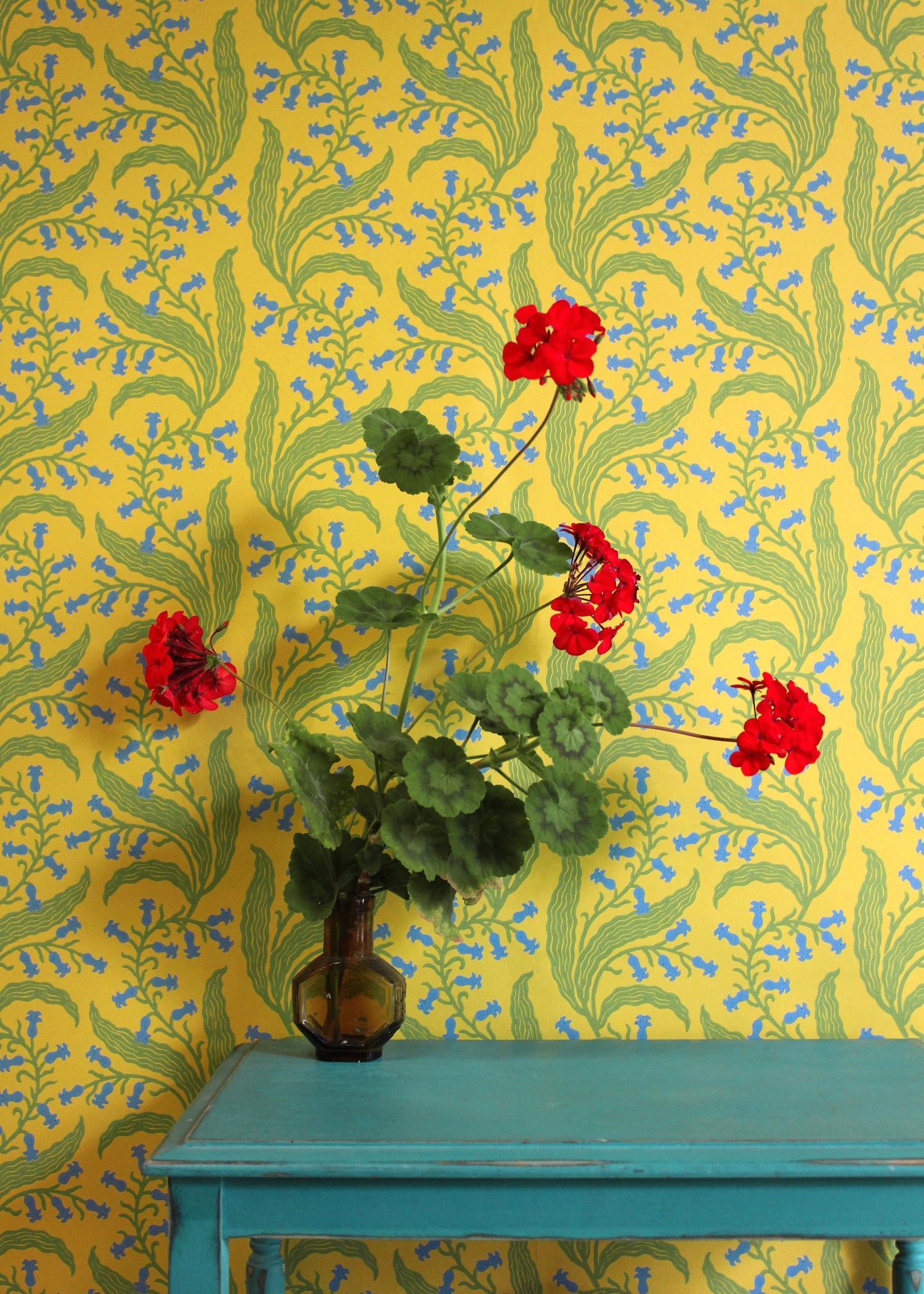 Teal table with vase and flowers against wallpaper in a scrolling floral pattern in blue and green on a yellow field. 