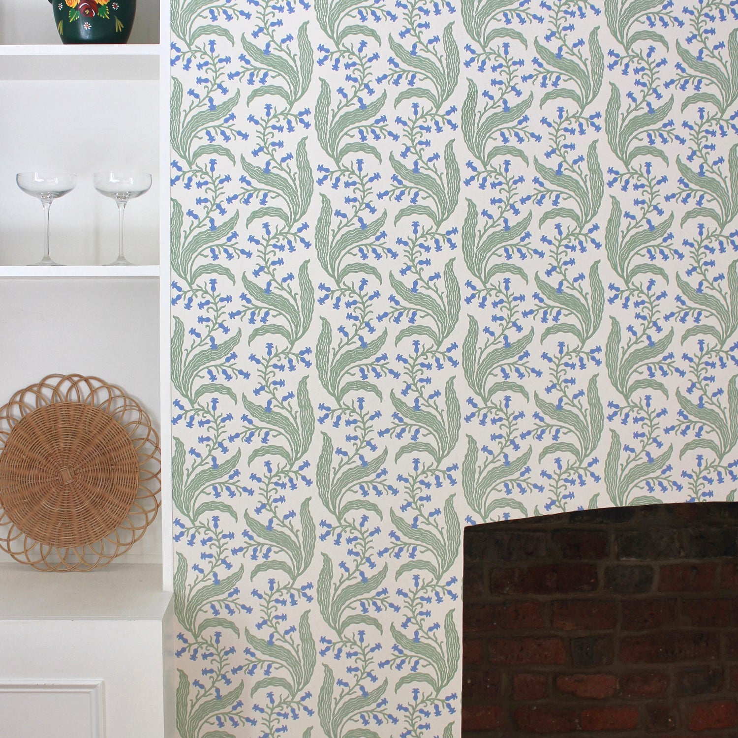 Wallpaper in a scrolling floral pattern in blue and green on a cream field surrounding a brick fireplace with a white bookshelf. 