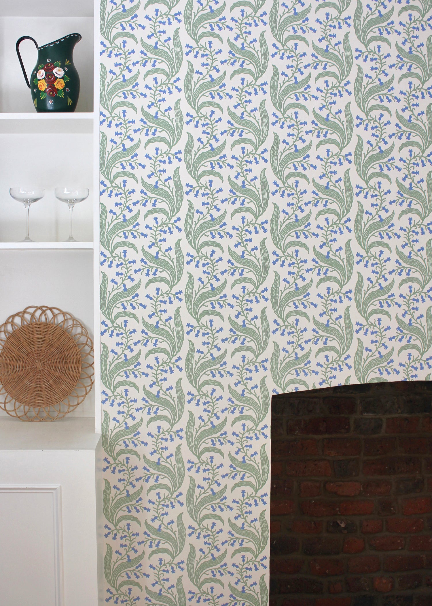 Wallpaper in a scrolling floral pattern in blue and green on a cream field surrounding a brick fireplace with a white bookshelf. 
